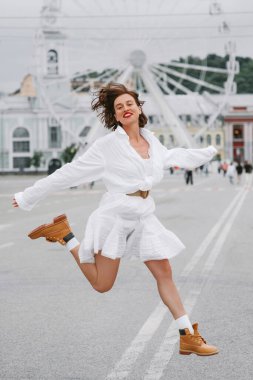 Güzel, mutlu bir kadın. Beyaz elbiseli, arka planda dönme dolap olan şehir meydanında zıplıyor.. 