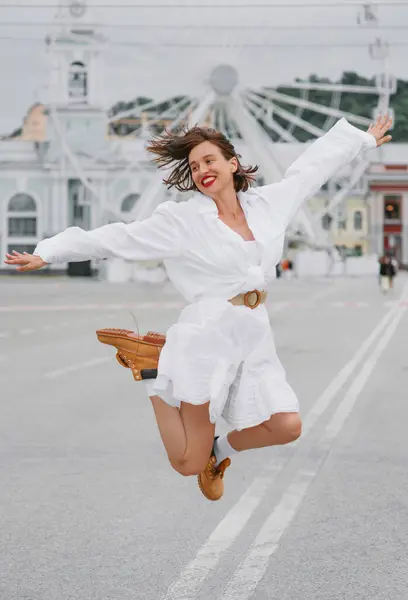 Güzel, mutlu bir kadın. Beyaz elbiseli, arka planda dönme dolap olan şehir meydanında zıplıyor.. 