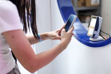 Bir kadın kasiyerle ödeme yapmak için akıllı telefonunu kullanır. Kart okuyucu kullanarak QR kodunu tarıyor ve daha az ödeme yaparak daha uygun bir bağlantı kuruyor..