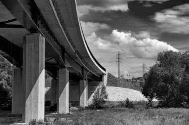 Arka planda bulutlu gökyüzü ve elektrik direkleriyle vadideki yol köprüsünün alt manzarası. Bir yol köprüsü ve siyah beyaz bir manzara..