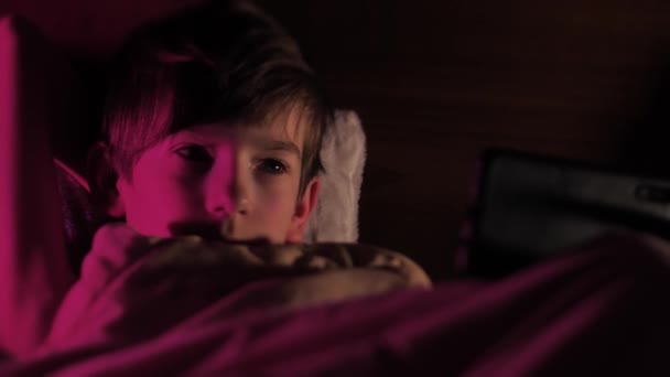 携帯電話を手に毛布の下のベッドの子供 小さな男の子がベッドでスマートフォンでビデオを見ています 子供は感情的に電話を見る 選択的ソフトフォーカス — ストック動画