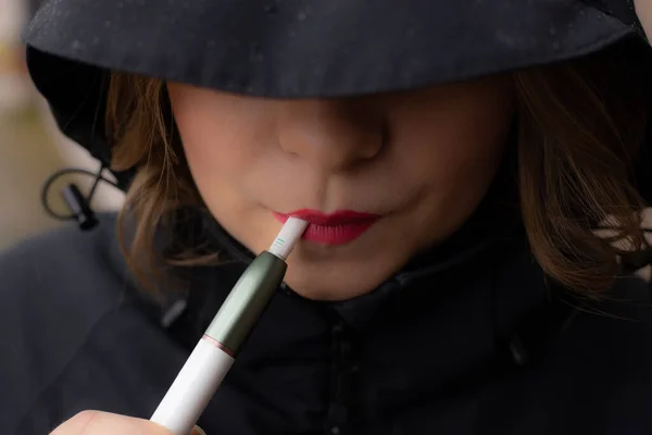 Kırmızı dudaklı bir kadın elektronik sigara içiyor. Kaputun altındaki güzel genç kadın. Güzel kırmızı dudaklı genç kız