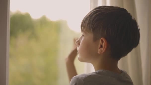 小さな男の子が窓のそばに立っている 少年は窓から手を振っている 家庭での育児 家で一人子 — ストック動画