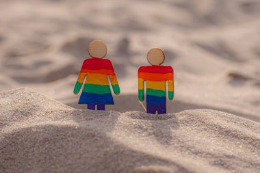 Kumsaldaki LGBT ahşap insanlar. Plajda duran çift, gökkuşağı bayrağıyla, eşit haklar, LGBT topluluğunun sembolü. Kumlu bir sahilde LGBT bayrağı. Yaz yolculuğu konsepti. Aynı cinsiyetin sembolü.