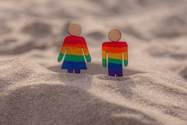 Kumsaldaki LGBT ahşap insanlar. Plajda duran çift, gökkuşağı bayrağıyla, eşit haklar, LGBT topluluğunun sembolü. Kumlu bir sahilde LGBT bayrağı. Yaz yolculuğu konsepti. Aynı cinsiyetin sembolü.