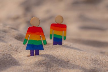 Kumsaldaki LGBT ahşap insanlar. Plajda duran çift, gökkuşağı bayrağıyla, eşit haklar, LGBT topluluğunun sembolü. Kumlu bir sahilde LGBT bayrağı. Yaz yolculuğu konsepti. Aynı cinsiyetin sembolü.