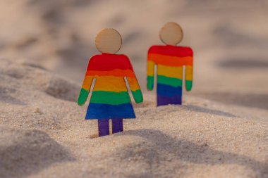 Kumsaldaki LGBT ahşap insanlar. Plajda duran çift, gökkuşağı bayrağıyla, eşit haklar, LGBT topluluğunun sembolü. Kumlu bir sahilde LGBT bayrağı. Yaz yolculuğu konsepti. Aynı cinsiyetin sembolü.