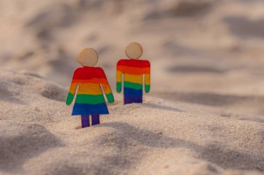 Kumsaldaki LGBT ahşap insanlar. Plajda duran çift, gökkuşağı bayrağıyla, eşit haklar, LGBT topluluğunun sembolü. Kumlu bir sahilde LGBT bayrağı. Yaz yolculuğu konsepti. Aynı cinsiyetin sembolü.