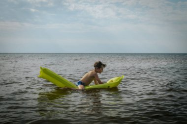 Denizde şişme yatakta yatan bir çocuk. Gülümseyen bir çocuk şişme bir yatakla denizde yüzüyor. Yumuşak seçici odak