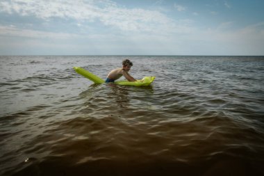 Denizde şişme yatakta yatan bir çocuk. Gülümseyen bir çocuk şişme bir yatakla denizde yüzüyor. Yumuşak seçici odak