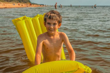 Denizde şişme yatakta yatan bir çocuk. Gülümseyen bir çocuk şişme bir yatakla denizde yüzüyor. Yumuşak seçici odak