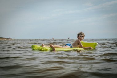 Denizde şişme yatakta yatan bir çocuk. Gülümseyen bir çocuk şişme bir yatakla denizde yüzüyor. Yumuşak seçici odak