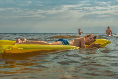 Denizde şişme yatakta yatan bir çocuk. Gülümseyen bir çocuk şişme bir yatakla denizde yüzüyor. Yumuşak seçici odak