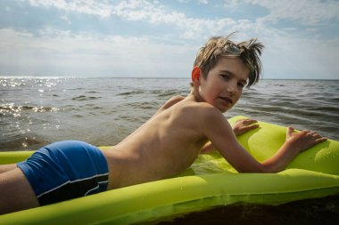 Denizde şişme yatakta yatan bir çocuk. Gülümseyen bir çocuk şişme bir yatakla denizde yüzüyor. Yumuşak seçici odak