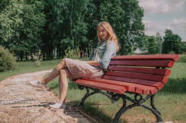 Genç bir kadın kırmızı bir bankta oturur ve kameraya bakar. Kız güzel doğanın tadını çıkarıyor. Yumuşak seçici odak