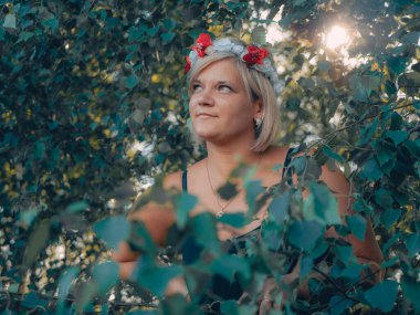 Çiçekli taç giymiş güzel bir kadın kamera için poz veriyor. Genç bir kadın huş dallarının arkasında saklanıyordu.