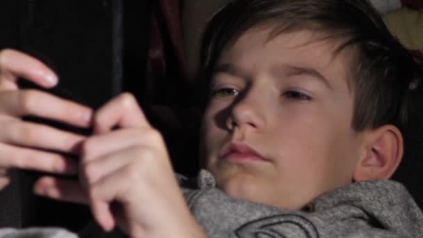 少年はベッドの上に横たわっている間に電話を見ている 携帯を持った子供が手元に — ストック動画