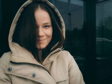 Krem rengi ceketli biri cam bir binanın önünde duruyor. Bireylerin yüzü gizlenmiş ve atmosfer soğuk ve modern görünüyor..