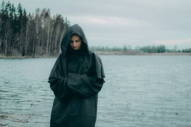 Siyah pelerinli bir kadın gölün kenarında duruyor.