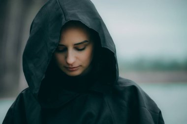 Siyah pelerinli bir kadın gölün kenarında duruyor.