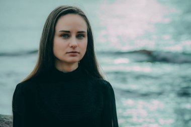 Koyu renk elbiseli bir kadın sahildeki bir kayanın üzerinde oturuyor.