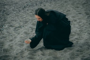 Deniz kenarında siyah elbiseli bir kadın bulanık doğal bir arka planda eline kum alır..