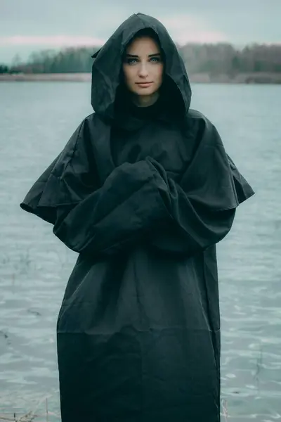 Siyah pelerinli bir kadın gölün kenarında duruyor.