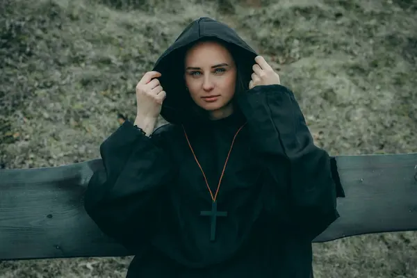 A woman in a black hoodie, hood up, face blurred for privacy, stands outdoors