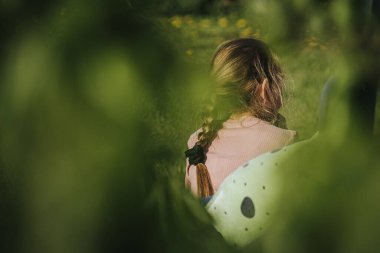 Arkadan görülen genç bir kız, saçları örgülü, sessizce bir bankta oturuyor, düşünceye dalmış..