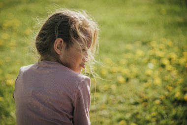 Genç bir kız, yüzü yumuşak güneş ışığıyla aydınlanmış, bulanık yeşil bir arka planda, omzunun üzerinden geriye bakar..