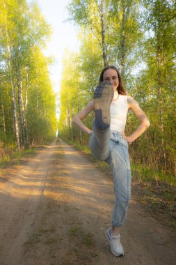 Neşeli bir kadın, uzun huş ağaçları ve yeşil yapraklarla kaplı güneşli bir orman yolunda yüksek bir tekme atar. Beyaz kolsuz bir bluz ve kot pantolon giyiyor. Enerjisini ve mutluluğunu gösteriyor.. 