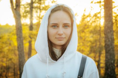 Parlak bir gülümsemesi ve yeşil gözleri olan beyaz kapüşonlu bir kadın, açık havada, arka planda güneşli ağaçlara ve altın yapraklara karşı duruyor. Görüntü mutluluk ve doğal güzelliği yakalar.