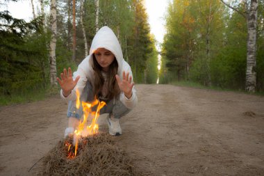 Beyaz kapüşonlu bir kadın orman yolunda çömeliyor, başlattığı küçük bir yangına odaklanıyor. Uzun huş ağaçları, yemyeşil yapraklarla yol boyunca uzanır, dingin ama yoğun bir şekilde.
