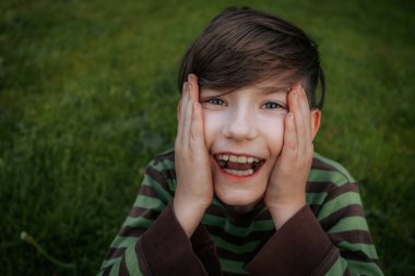 Çizgili gömlekli genç bir çocuk çimlerin üzerinde oturuyor, ağzı açık bir şekilde şaşkınlıkla yüzünü tutuyor. Yeşil arkaplan oyuncu ifadeye doğal, canlı bir ayar ekler.