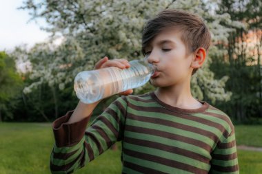 Plastik şişeden su içen, çizgili tişörtlü ve arka planda bulanık yeşil bir çocuğun yakın plan görüntüsü..