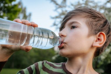 Plastik şişeden su içen, çizgili tişörtlü ve arka planda bulanık yeşil bir çocuğun yakın plan görüntüsü..