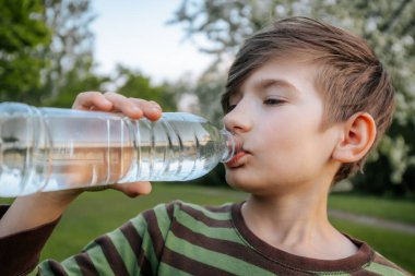 Plastik şişeden su içen, çizgili tişörtlü ve arka planda bulanık yeşil bir çocuğun yakın plan görüntüsü..