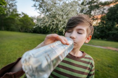 Plastik şişeden su içen, çizgili tişörtlü ve arka planda bulanık yeşil bir çocuğun yakın plan görüntüsü..