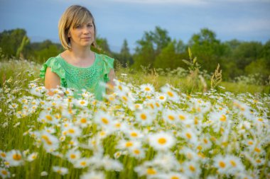 Yeşil gömlekli bir kadın sarı merkezli beyaz papatya tarlasında ellerini uzatıyor. Arka planda yemyeşil ve berrak bir gökyüzü var. Huzurlu ve huzurlu bir hava yaratıyor..
