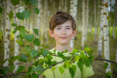 Açık yeşil gömlek giyen genç bir çocuk huş ağacının ormanında yapraklı bir dalı tutarken gülümsüyor. Arka planda, sakin ve doğal bir ortam oluşturan huş ağaçları sıraları yer alıyor..