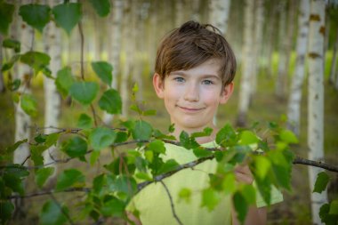 Açık yeşil gömlek giyen genç bir çocuk huş ağacının ormanında yapraklı bir dalı tutarken gülümsüyor. Arka planda, sakin ve doğal bir ortam oluşturan huş ağaçları sıraları yer alıyor..