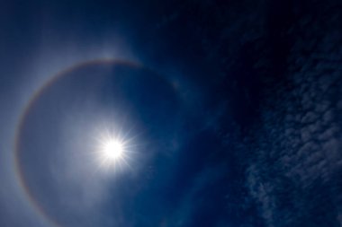 Güneş halesinin, atmosferdeki buz kristallerinin arasından geçen güneş ışığının kırılmasıyla oluşan derin mavi gökyüzüne karşı büyüleyici bir görüntüsü. Güneş parlak, dairesel gökkuşağı gibi bir hale ile çevrili..