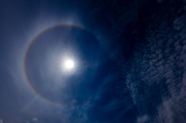 Güneş halesinin, atmosferdeki buz kristallerinin arasından geçen güneş ışığının kırılmasıyla oluşan derin mavi gökyüzüne karşı büyüleyici bir görüntüsü. Güneş parlak, dairesel gökkuşağı gibi bir hale ile çevrili..
