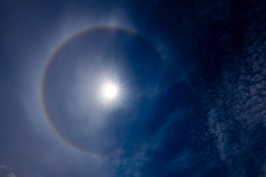 Güneş halesinin, atmosferdeki buz kristallerinin arasından geçen güneş ışığının kırılmasıyla oluşan derin mavi gökyüzüne karşı büyüleyici bir görüntüsü. Güneş parlak, dairesel gökkuşağı gibi bir hale ile çevrili..