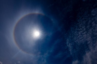 Güneş halesinin, atmosferdeki buz kristallerinin arasından geçen güneş ışığının kırılmasıyla oluşan derin mavi gökyüzüne karşı büyüleyici bir görüntüsü. Güneş parlak, dairesel gökkuşağı gibi bir hale ile çevrili..