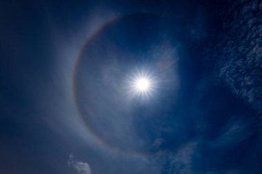 Güneş halesinin, atmosferdeki buz kristallerinin arasından geçen güneş ışığının kırılmasıyla oluşan derin mavi gökyüzüne karşı büyüleyici bir görüntüsü. Güneş parlak, dairesel gökkuşağı gibi bir hale ile çevrili..