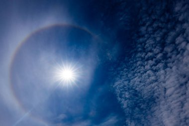 Güneş halesinin, atmosferdeki buz kristallerinin arasından geçen güneş ışığının kırılmasıyla oluşan derin mavi gökyüzüne karşı büyüleyici bir görüntüsü. Güneş parlak, dairesel gökkuşağı gibi bir hale ile çevrili..