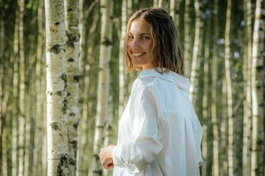 Bir huş ağacı ormanında dururken kısa saçlı bir kadın sıcak bir şekilde gülümsüyor. Beyaz bir gömlek giyiyor ve güneş ışığı ağaçların arasından süzülüp neşeli ve doğal bir atmosfer yaratıyor..