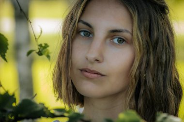 Yeşil gözlü, doğal ve sakin bir yüz ifadesi olan bir kadının yakın çekimi. Arka plan bulanık yeşillik gösteriyor, yüz hatlarına odaklanmasını sağlıyor..