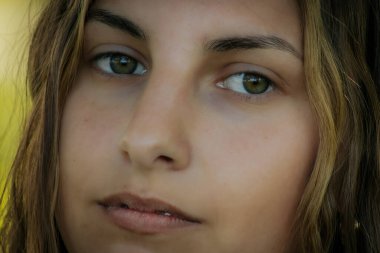 Yeşil gözlü, doğal ve sakin bir yüz ifadesi olan bir kadının yakın çekimi. Arka plan bulanık yeşillik gösteriyor, yüz hatlarına odaklanmasını sağlıyor..